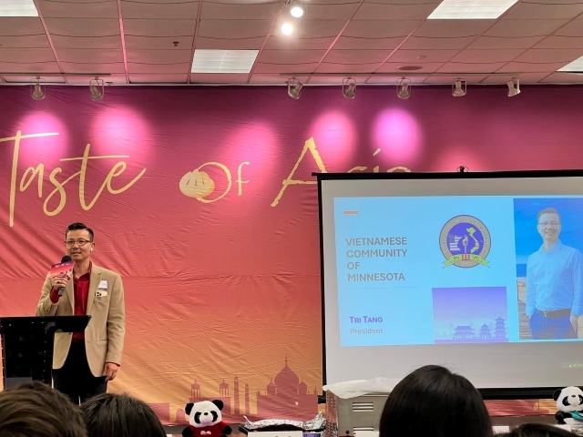 A man with a microphone speaks to a crowd in front of a pink and yellow screen, with a second screen and a projected image in front of it.
